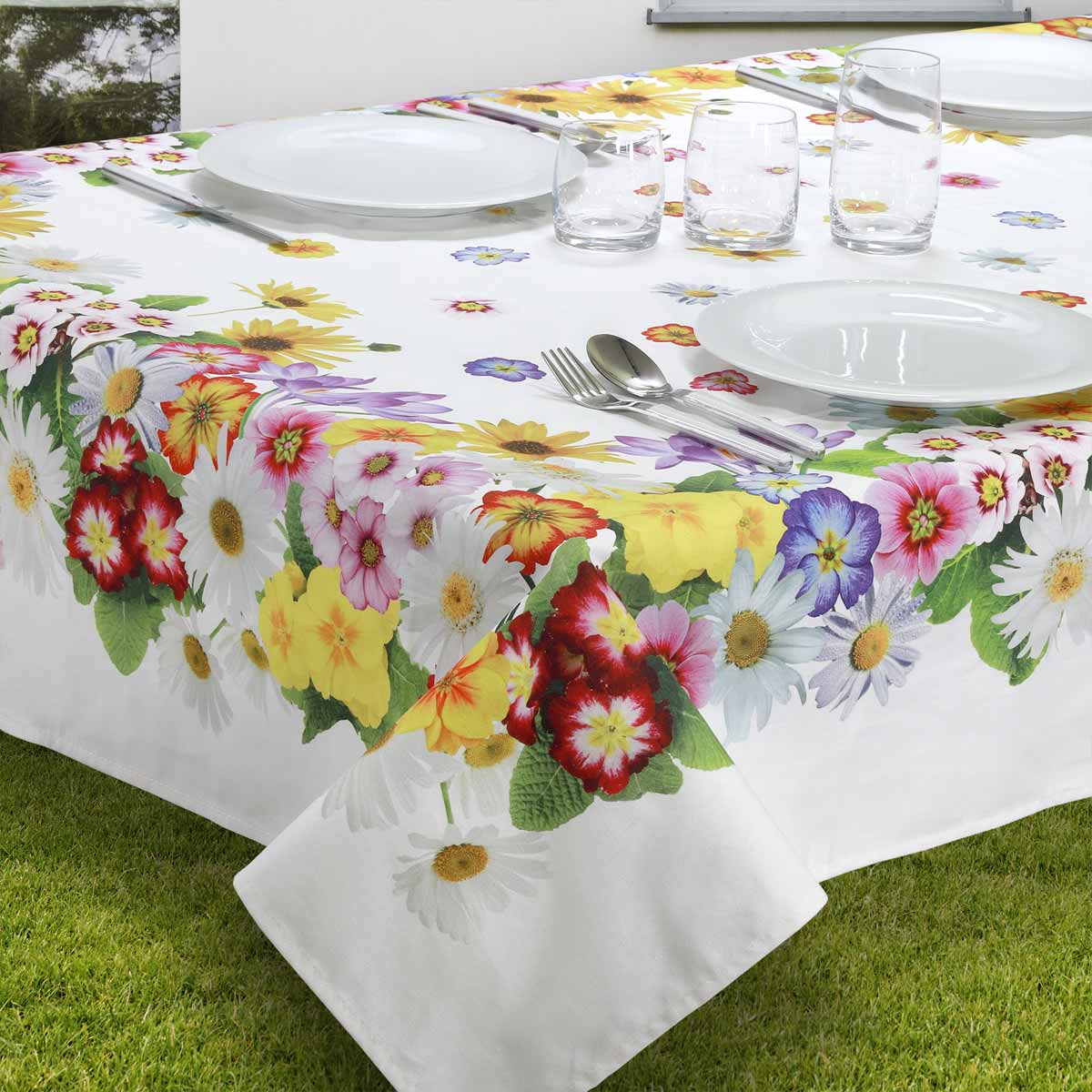 Tovaglia bianca in puro cotone su un tavolo apparecchiato all'aperto, decorata con una vivace bordatura di fiori colorati tra cui margherite, girasoli e primule.